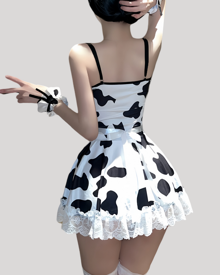 Woman wearing a cow-print maid costume set, featuring a lace-trimmed bodice, white apron with black bow, matching frilled headband and wrist cuffs, and sheer thigh-high stockings.