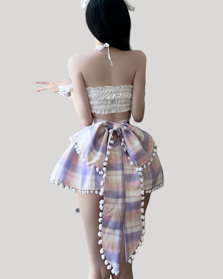 Woman wearing a pastel plaid bunny costume set, featuring a white bandeau top with pom-pom bows, a pleated mini skirt trimmed with white pom-pom fringe, matching plaid bunny ears headband, and ruffled wrist cuffs.